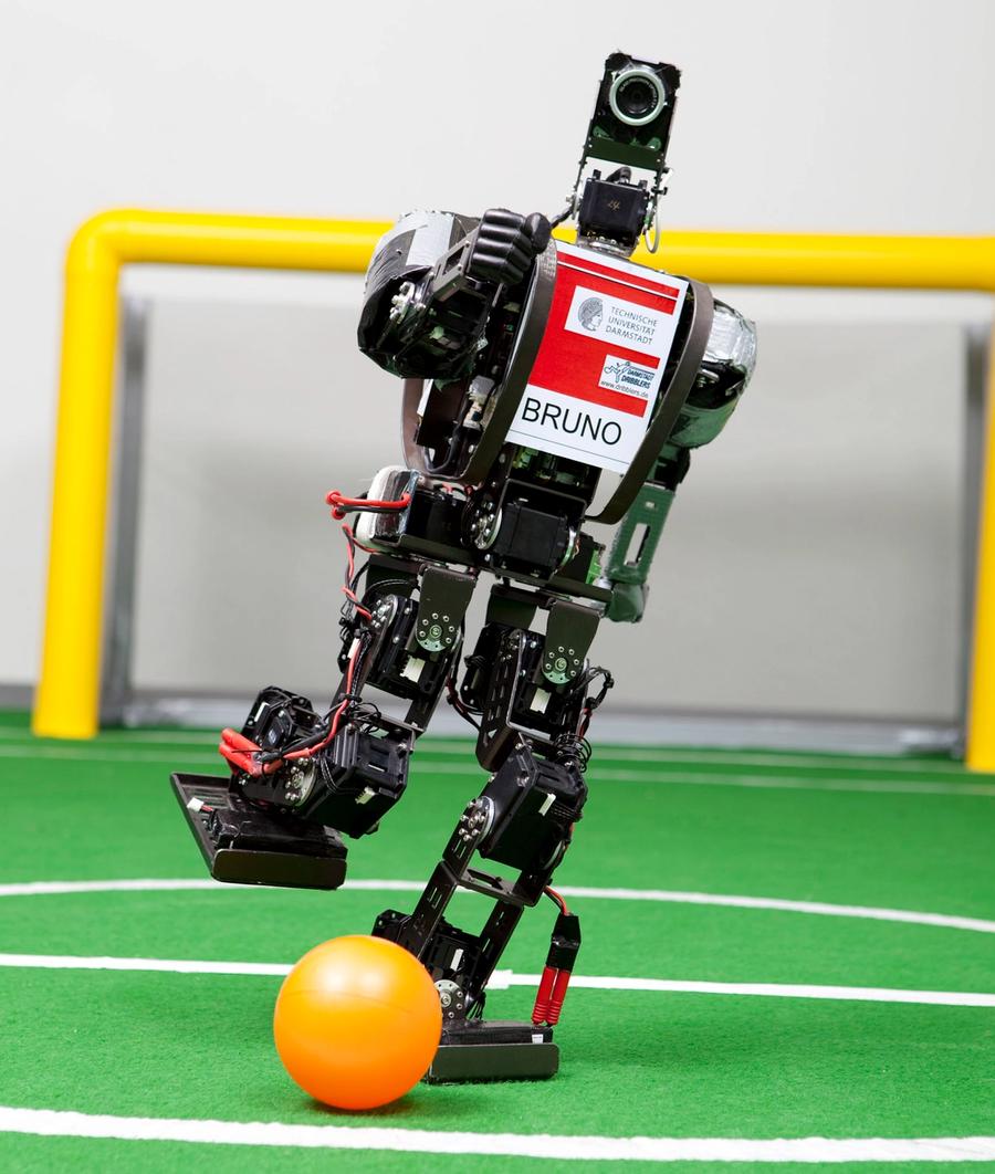 A black two-legged robot labelled Bruno kicks a orange ball on a miniature soccer field.