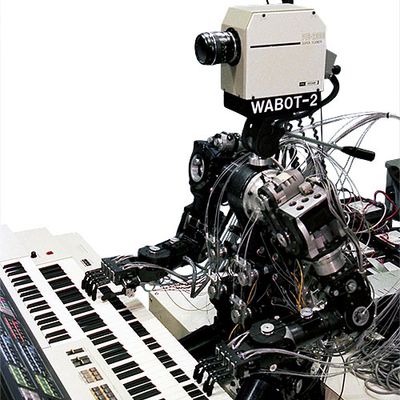 A historical robot with a complex humanoid body and a 1980's era video camera for a head sits and plays the keyboards.