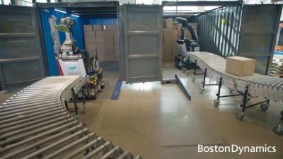 Two Stretch robots, which are large machines with a mobile wheeled base and a big articulated manipulator arm with suction cups, pick up cardboard boxes from inside shipping containers and place them onto a conveyor belt at Boston Dynamics' test facility.