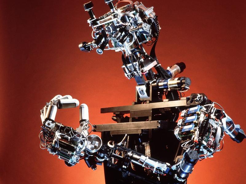 A human shaped robot with exposed electronics looks at it's fingers, which are pushed together to form an O.