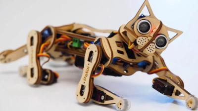 Small robot cat made of laser-cut wood, electronics, and wires rests on a while desk.