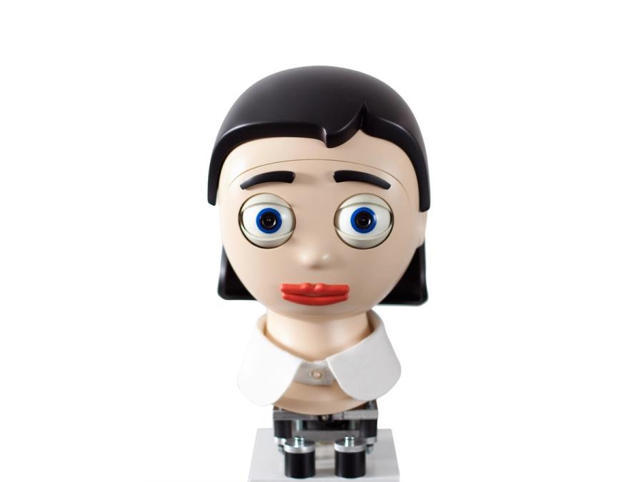 A cartoonish robotic head with big blue eyes with cameras, red lips, and black eyebrows and a black, bobbed hairdo sits on a base.