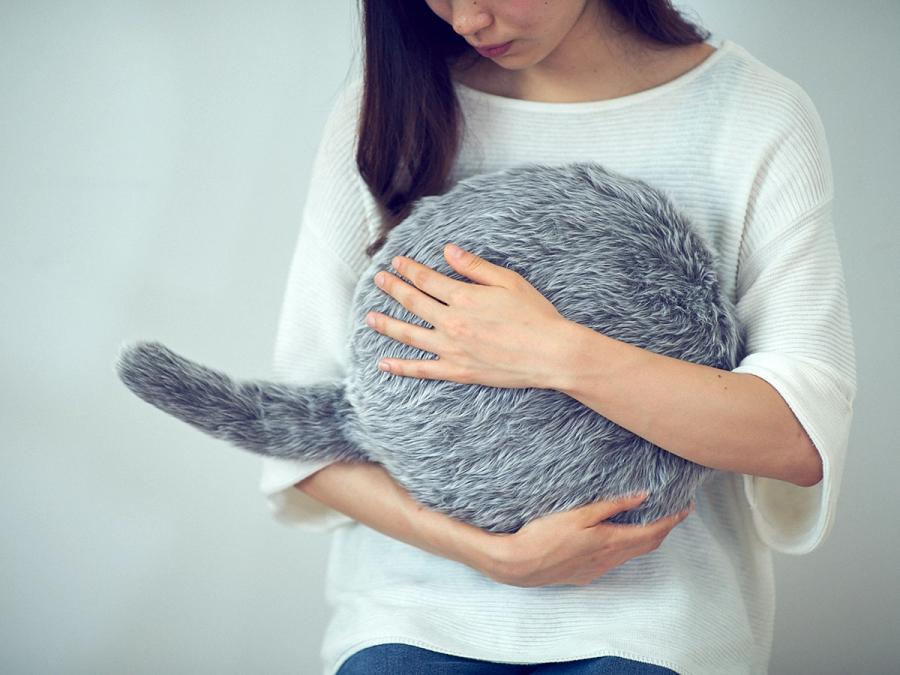 A dark haired woman in a white shirt cuddles a round cushion with a tail, covered in soft looking gray fur, to her chest.