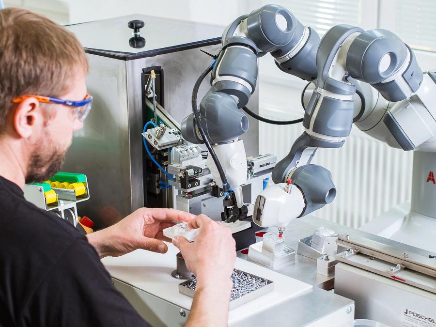 A white base and torso with two industrial arms works with a person wearing safety glasses, and holding plastic piece in his hands, in a factory setting.