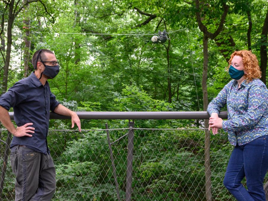 Two people wearing masks lean on a fence outdoors. In the distance behind them, the SlothBot robot hangs from a wire.