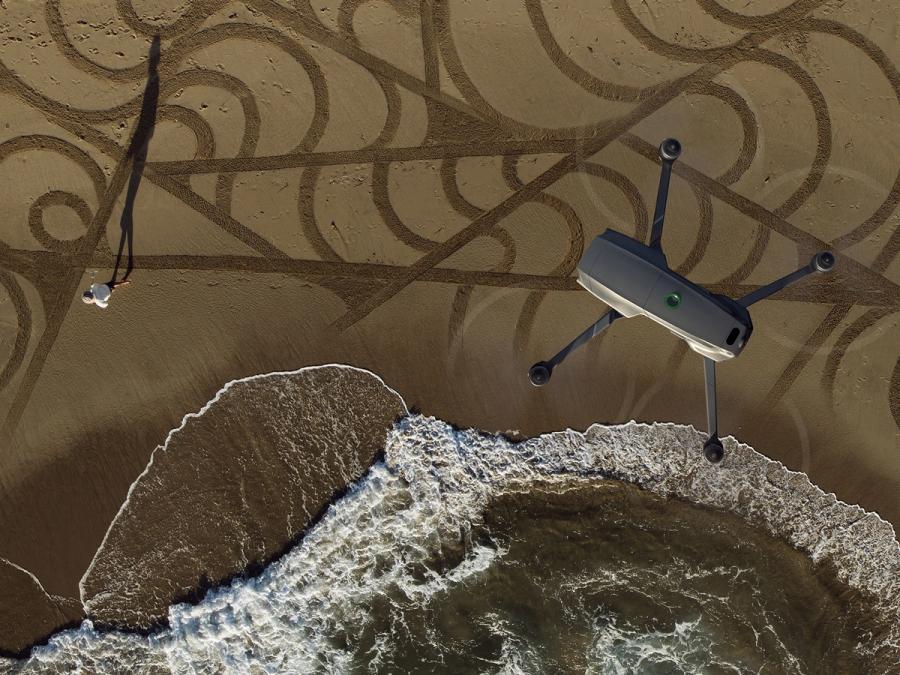 An aerial photo shows the drone in the air above a sandy beach with water and a person walking.