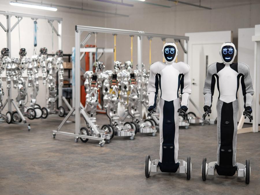 Two white and black EVE humanoid robots on wheeled bases, with gripper arms, and display faces showing round eyes and smiling mouths, stand in front of rows of humanoid body structures at the 1X Technologies company.