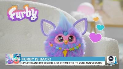 A purple Furby sits on a white couch, with overlay text that says in part Furby is back.