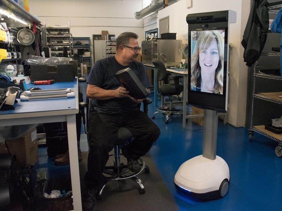 A man in a workspace holds an item up to show a woman displayed on a telepresence robot.