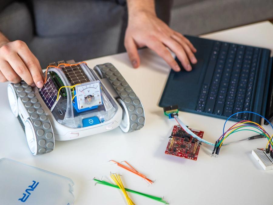 A small mobile vehicle with a base, tracked wheels and electronics sits on a desk next to a laptop.