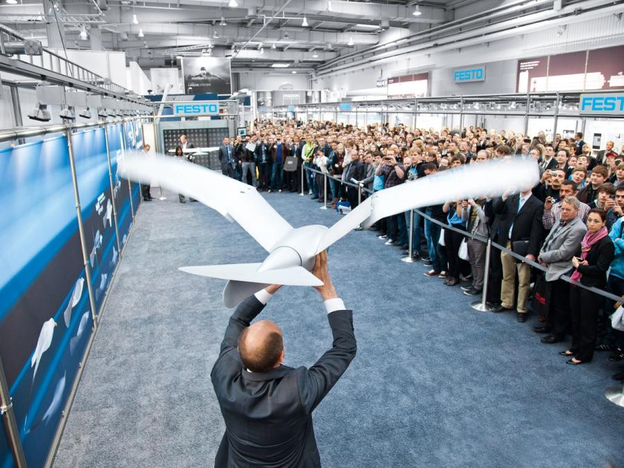 A man in a suit prepares to throw the robotic seagull, by holding it over his head. The robot's torso alone is larger then his head. A large crowd is watching, but cordoned off.