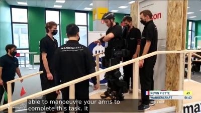 A man wearing a pair of robotic legs prepares to walk down a wooden ramp during an assistive robotics technology competition, while five other people stand nearby observing.