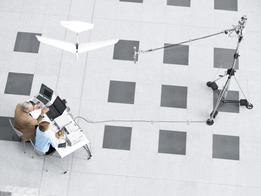 Aerial photo shows two people sitting at a desk with computers while the SmartBird flies overhead, indoors.