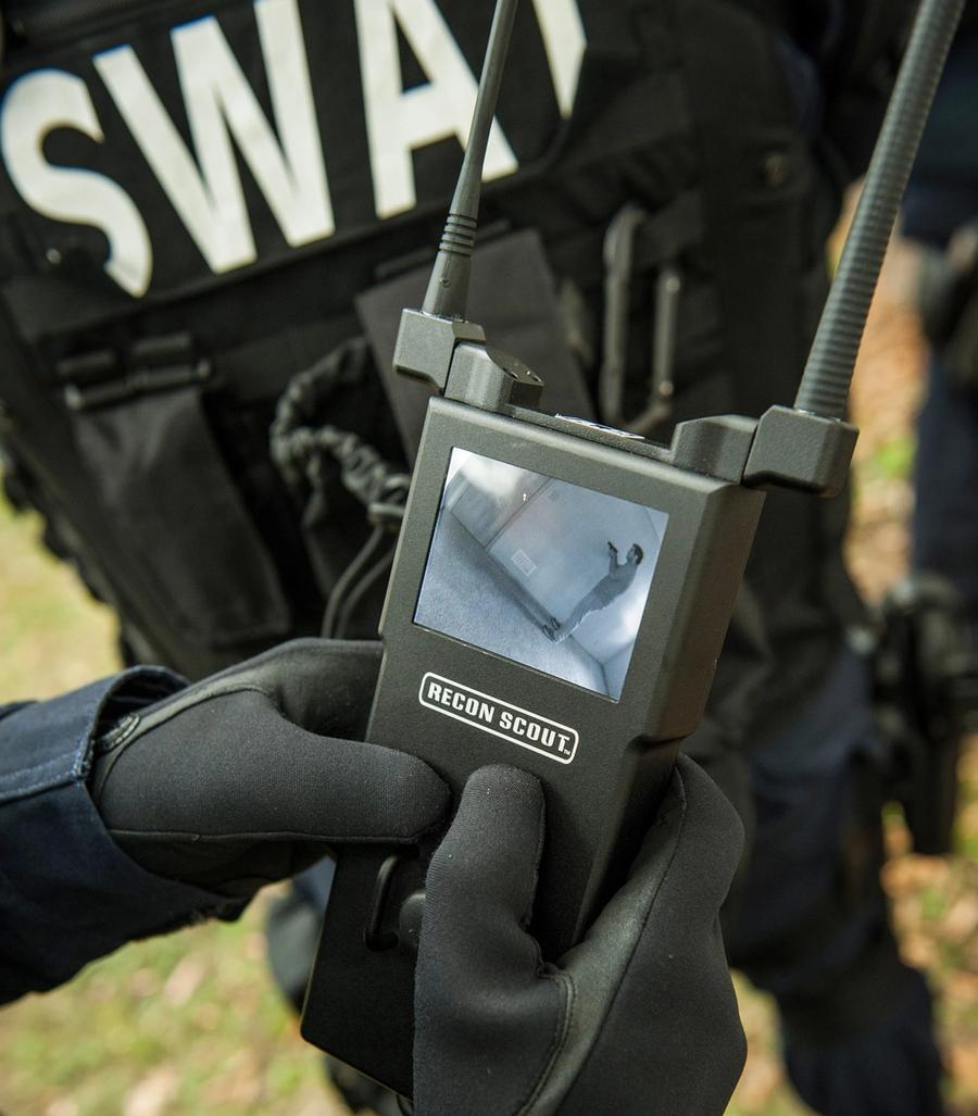 Close up of gloved hands holding a device that looks like a walkie talkie, but has a display on it which shows the robots perspective.