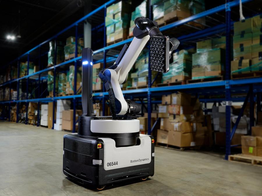 Stretch, a large black and grey mobile base with an articulated arm ending in a rectangular suction pad, sits in a factory. A vertical black column with a light extends from the base.