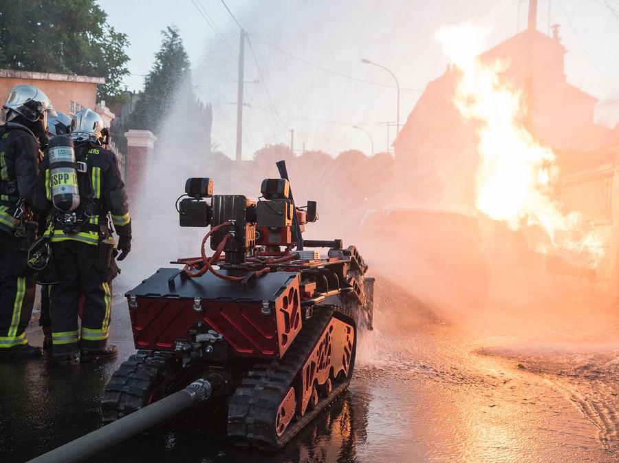 A red robotic vehicle the size of a small tank blasts water on a fire while two human firefighters observe.