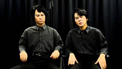 An android that looks just like Japanese researcher Hiroshi Ishiguro sits next to the real Hiroshi Ishiguro, who has a wide face, dark hair, furrowed eyebrows, and wears black shirt and pants.