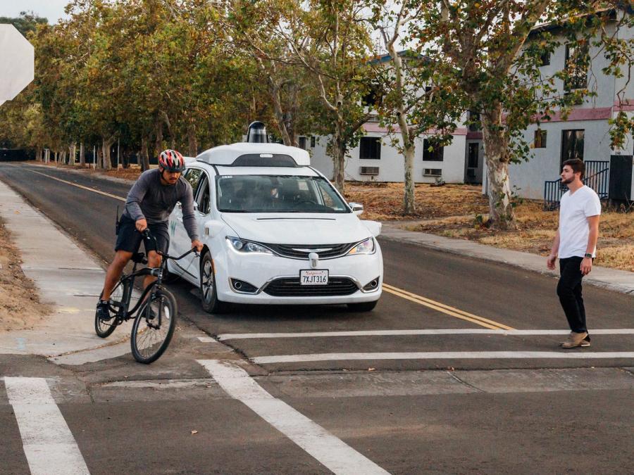 A white car with a small black dome on its top in a city road scene with a biker on its left and man walking in a crosswalk in front of it.