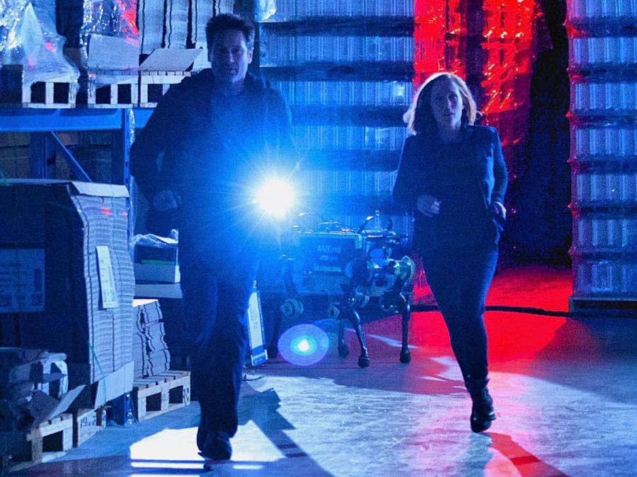 A running woman and man with a flashlight in a dark blue and red lit building. The robot is behind them.