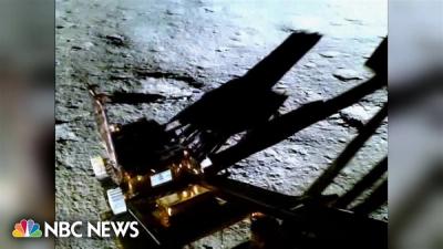 The Pragyan rover, with silver metal wheels and golden rectangular body, rolls down a ramp, casting a shadow on the gray lunar surface.