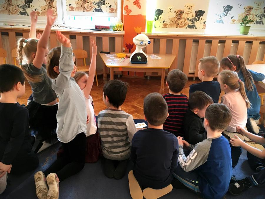 The robot sits on a table in front of a group of children seated on a rug.