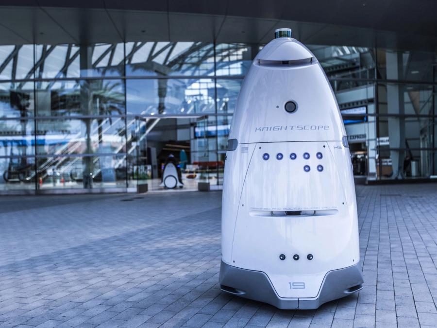 A white, rocket shaped rolling robot stands outside a building.