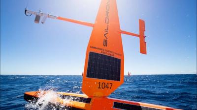 A Saildrone, which looks like a boat with a red vessel, floats in a dark blue ocean against a bright sunny cloudless blue sky.