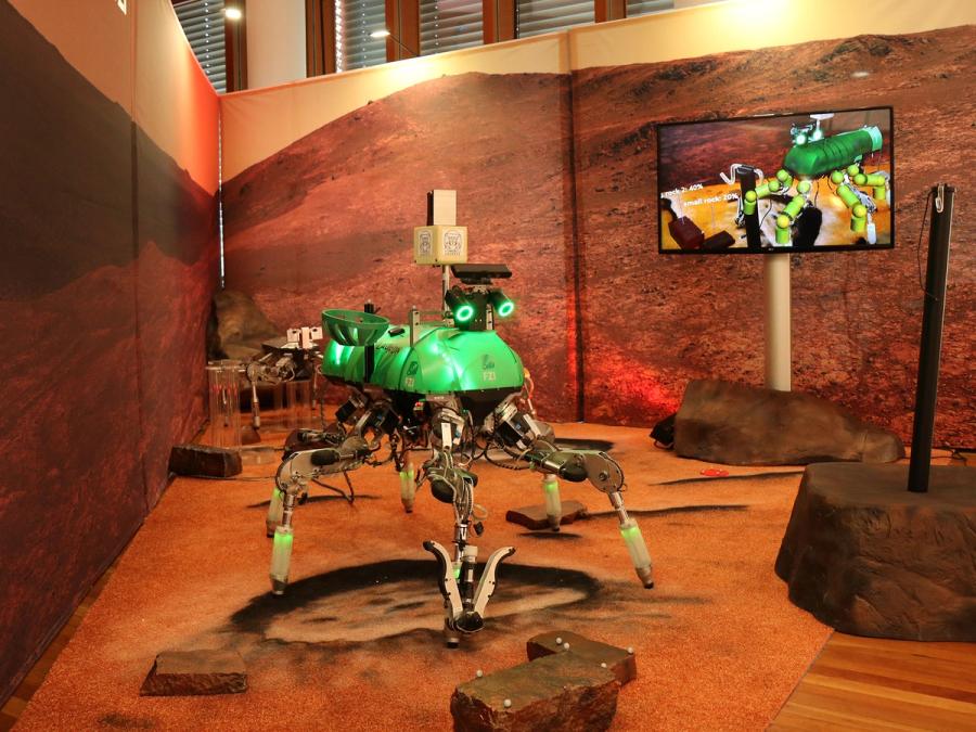 The hexapod robot walks on a red martian- like surface. Its movements are displayed on a monitor behind it.