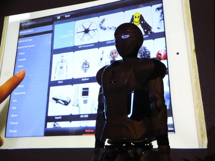 An adult-size humanoid robot is seen in a semi-dark room with a screen showing other robots in the background.