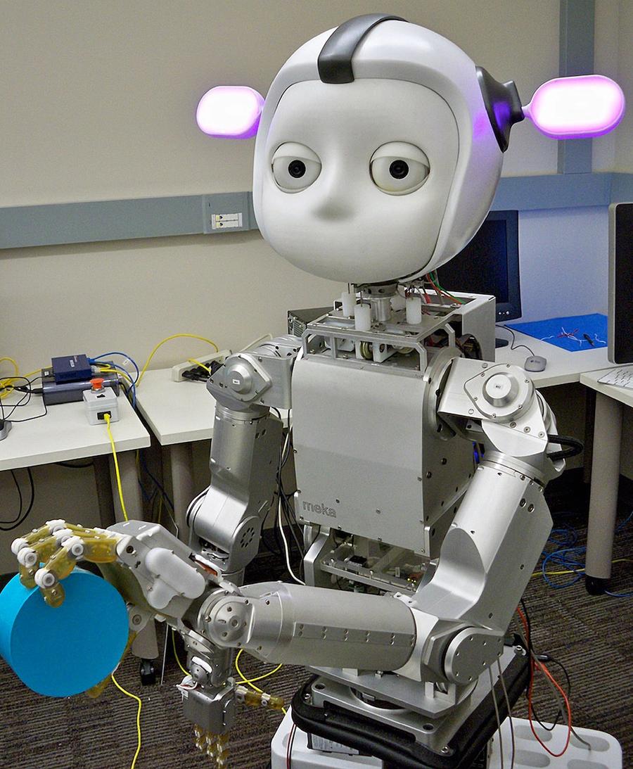 A robot with a metal body, round face with big eyes and pink ears, holds a blue object and looks at the camera, with a lab in the background.