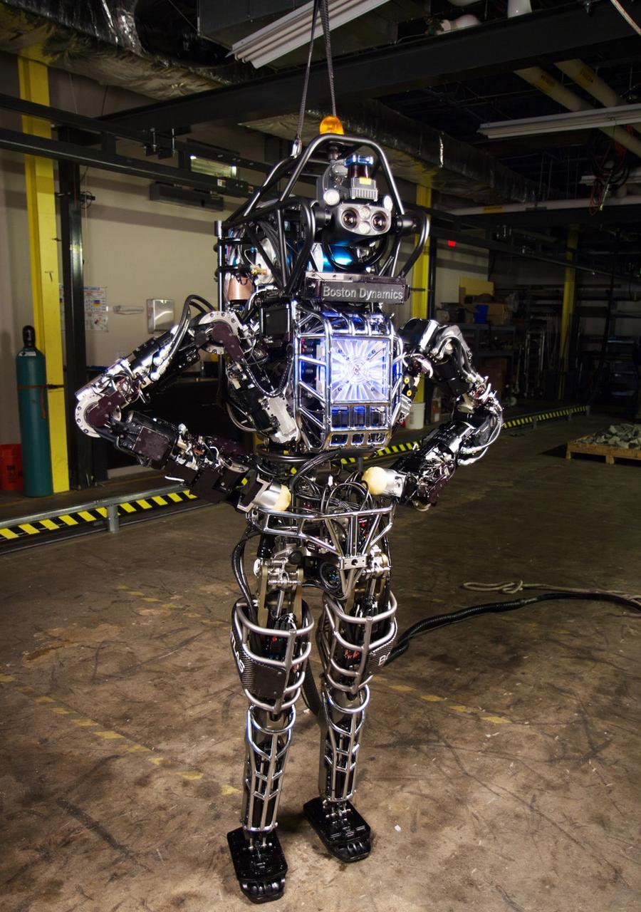 A two legged robot named Atlas 2013 with its hands on hips poses in a warehouse.