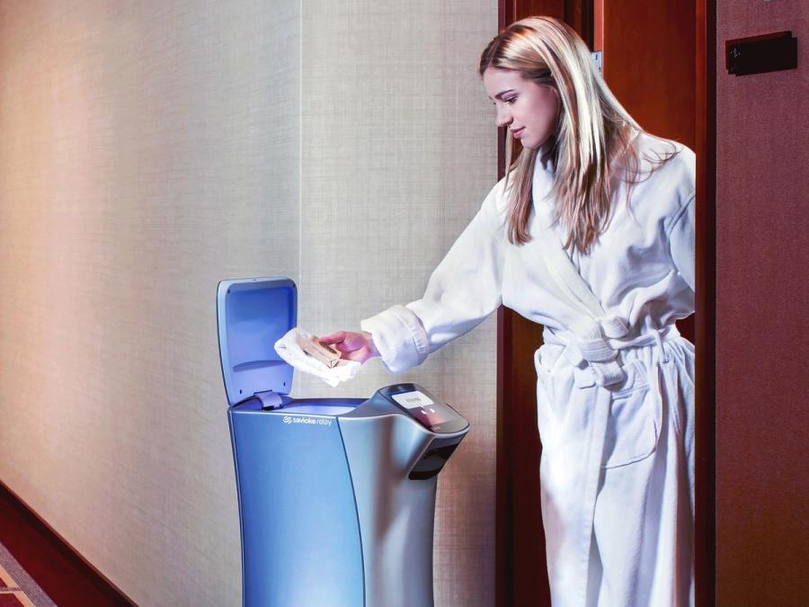 View of the top of the robot where its top compartment lid is open and delivering something a woman in the door of a hotel room.