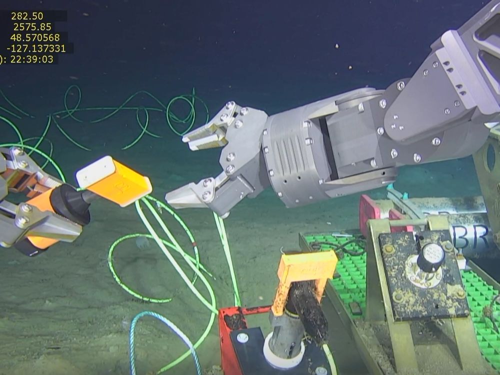 Near the ocean floor, a robotic claw reaches for a tool that another robotic claw is grasping.