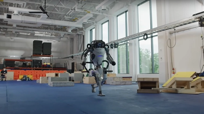 Atlas, a adult-sized metal and plastic humanoid robot, runs inside a lab with blue padded floors and large glass windows.