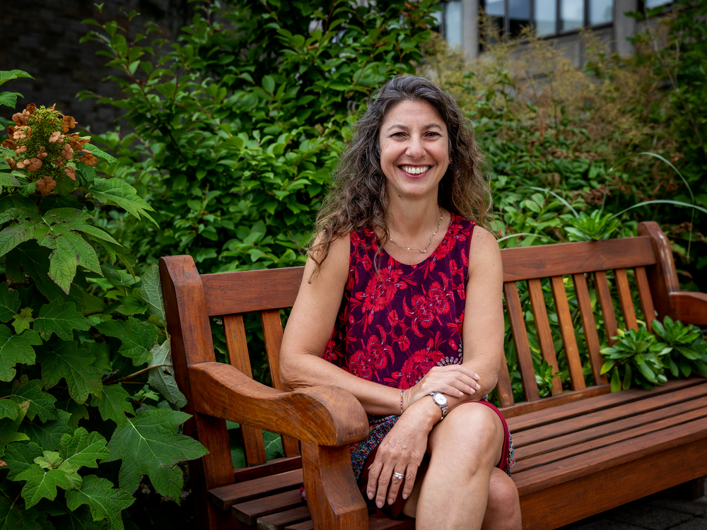 A photo of a smiling woman sitting on a bench in front of plants.  