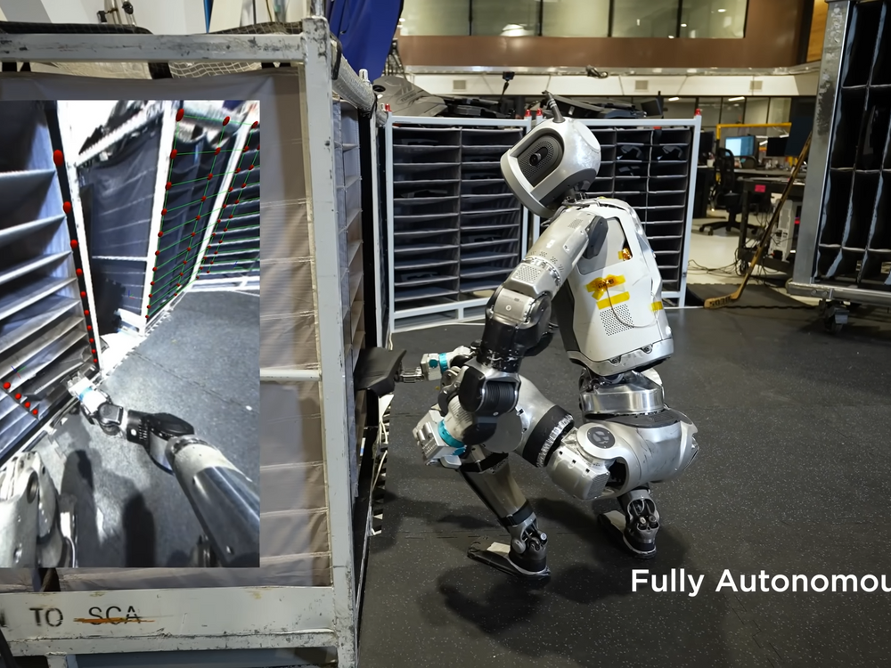 A silvery humanoid robot squats down to grasp a car part out of a bin in a research environment.